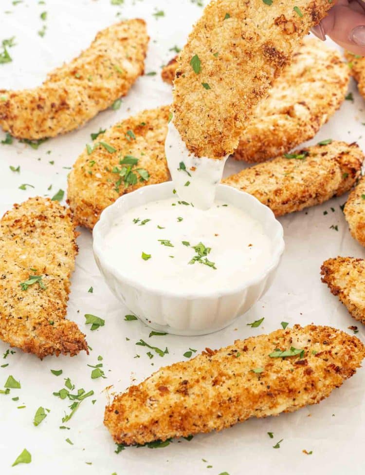 Hand dipping a golden air fryer chicken tender into ranch dressing, surrounded by more tenders and herbs.