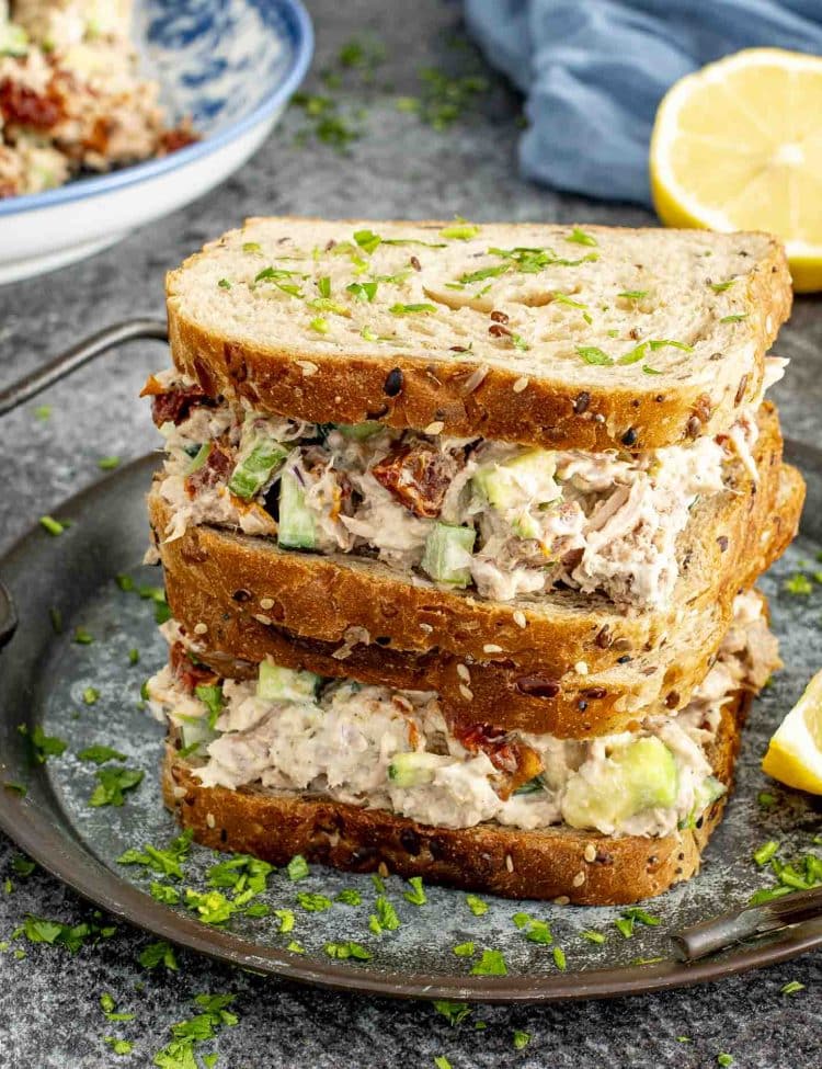 Hearty avocado tuna salad sandwich stacked high with cucumbers and sun-dried tomatoes on multigrain bread.