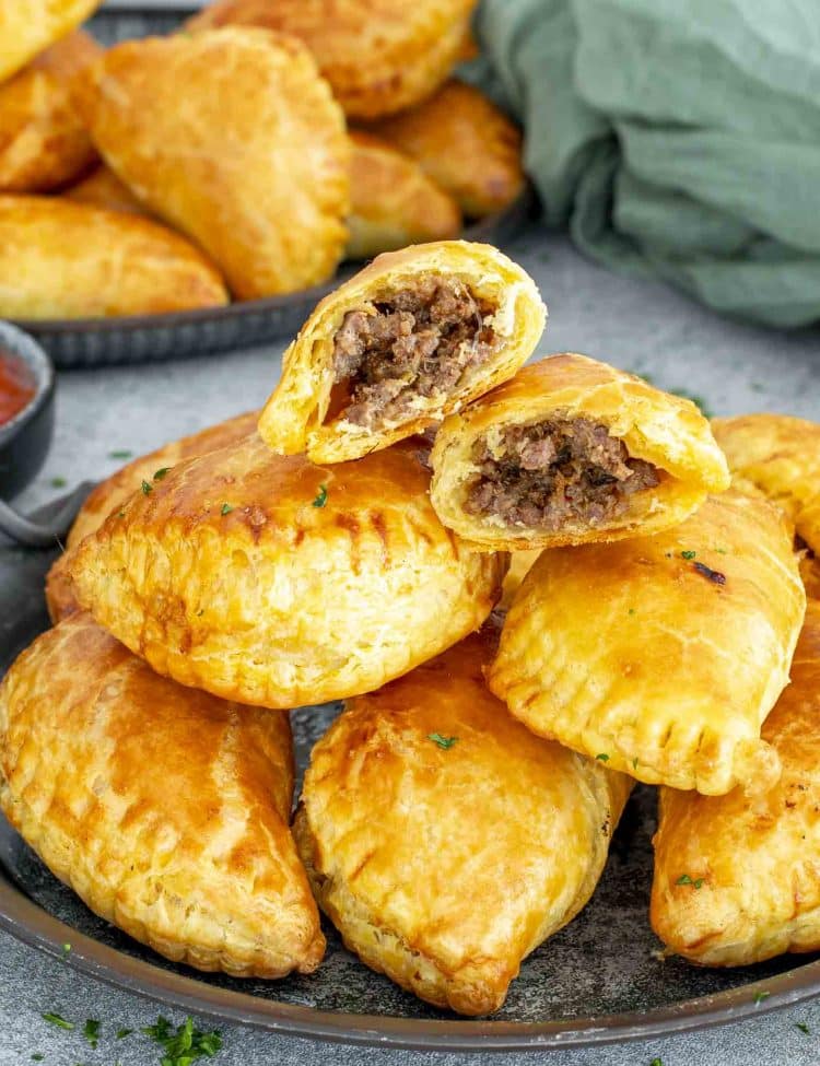 Stack of beef empanadas on a platter, with one empanada cut open revealing juicy meat filling.
