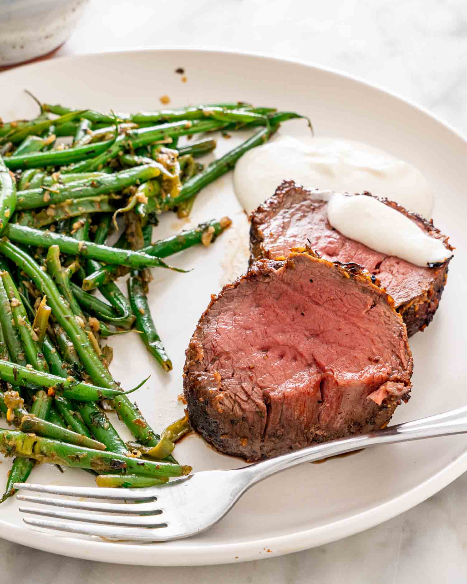 2 slices of beef tenderloin with skillet green beans and horseradish yogurt sauce on a white plate.