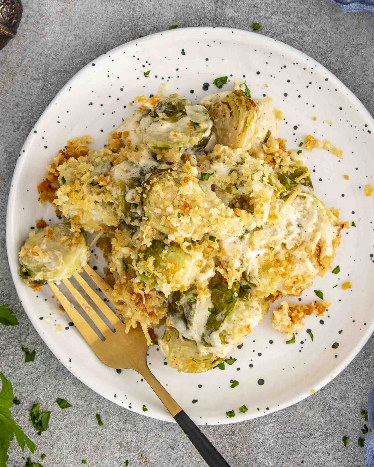 Plate of creamy Brussels sprouts gratin topped with Gruy&egrave;re, Parmesan, and crispy breadcrumbs, served with gold fork and parsley garnish.