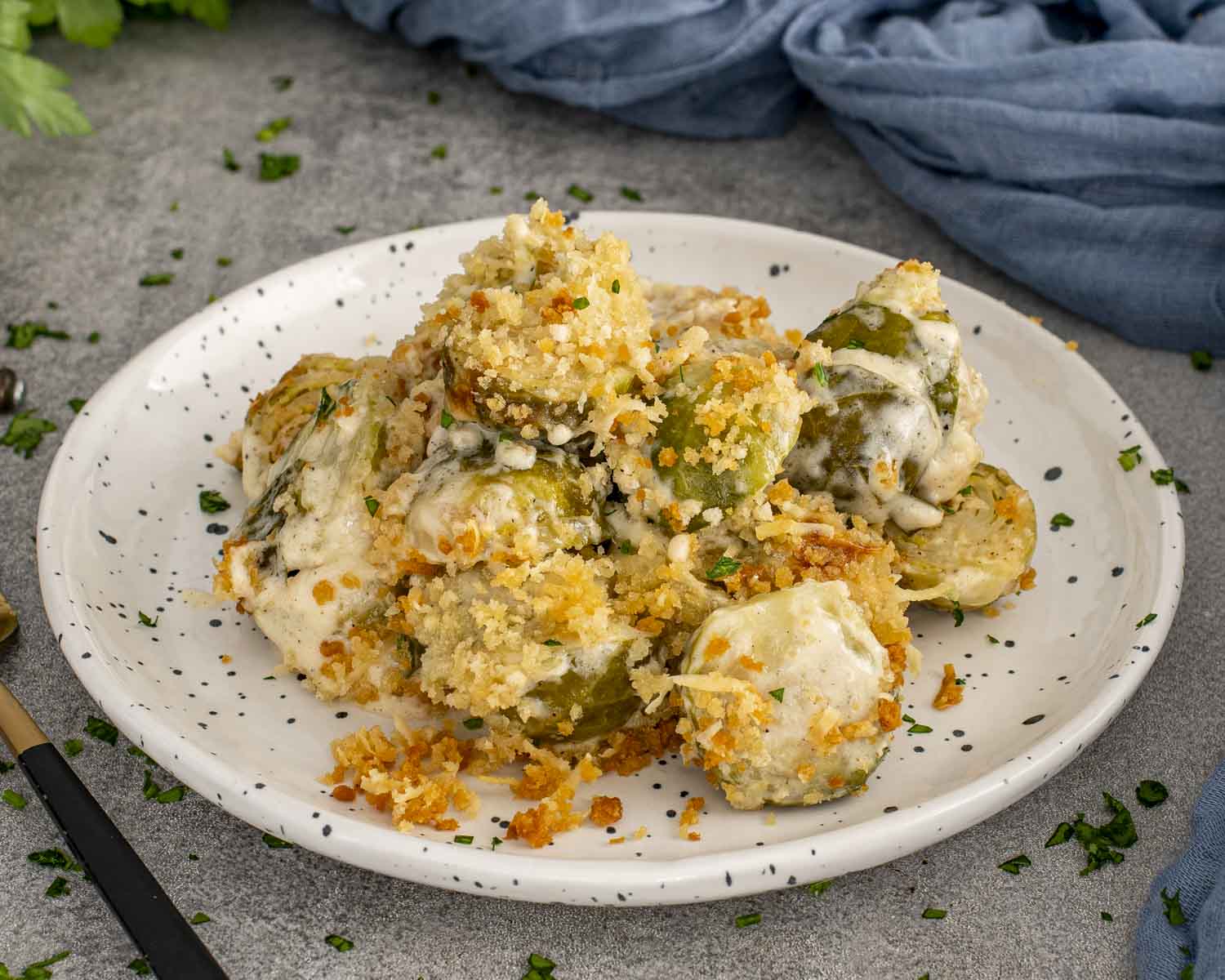 Cheesy Brussels sprouts gratin piled on a white speckled plate, topped with golden panko crumbs and fresh parsley.