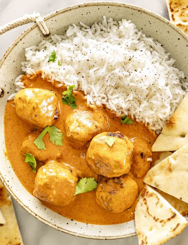overhead shot of butter chicken meatballs with a side of rice in a bowl with naan slices and garnished with cilantro.