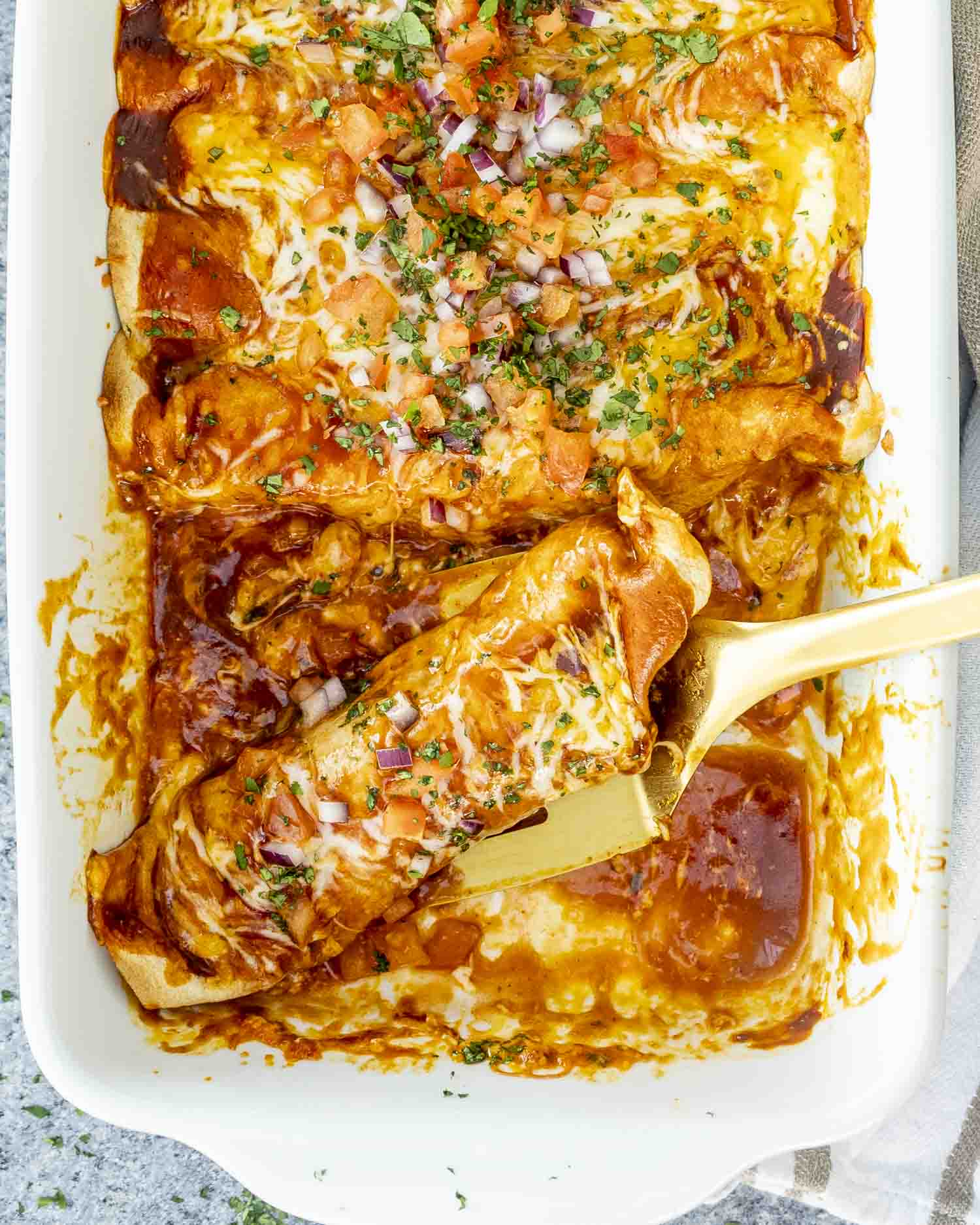 A golden spatula lifting enchiladas from a half-empty baking dish with melted cheese and sauce coating.
