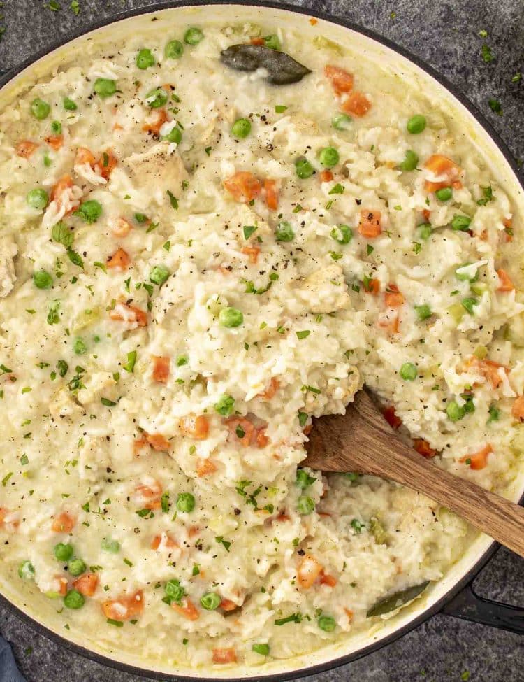 Large skillet filled with creamy chicken rice pilaf, flecked with peas, carrots, and cracked black pepper.