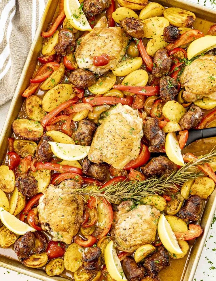 Sheet pan loaded with baked chicken, sausage, potatoes, peppers, lemon wedges, and fresh rosemary sprig.