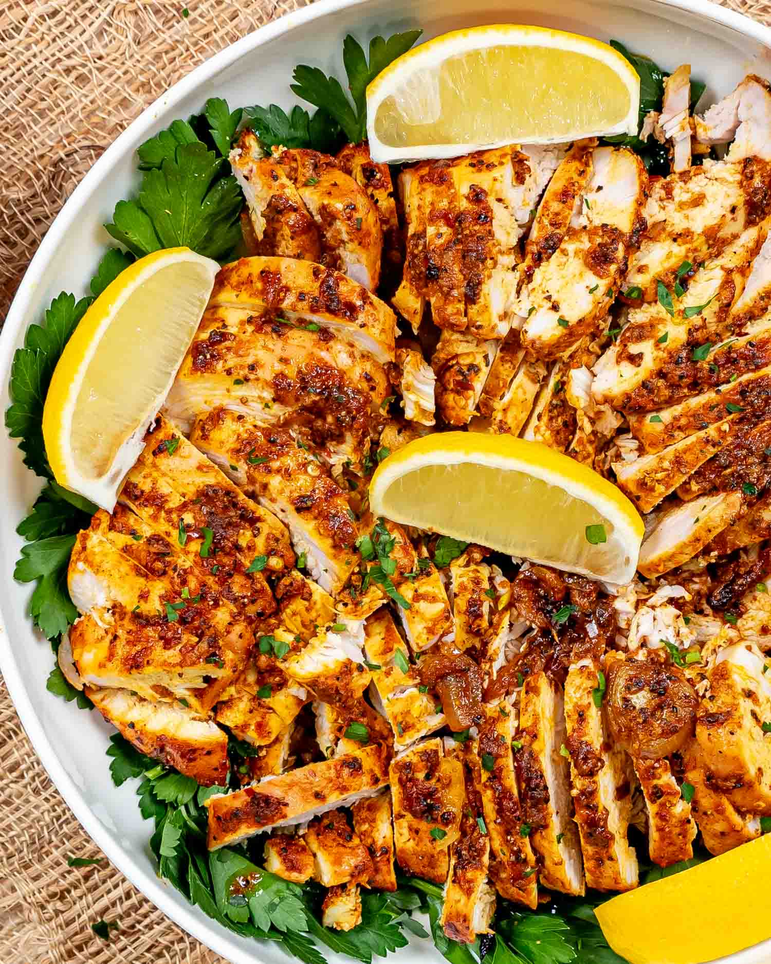 sliced chicken shawarma in a white bowl garnished with parsley and lemon wedges.