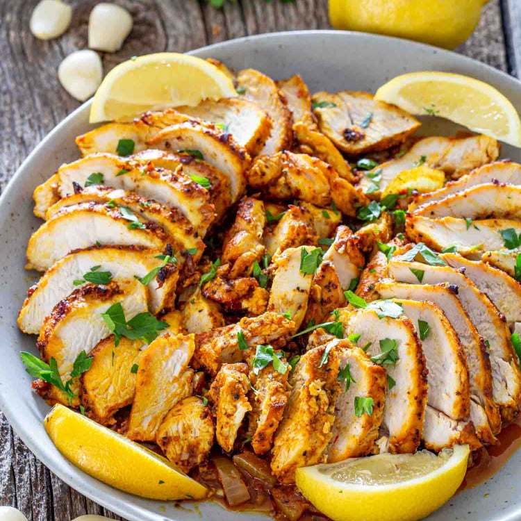 sliced chicken shawarma in a white bowl garnished with parsley and lemon wedges.