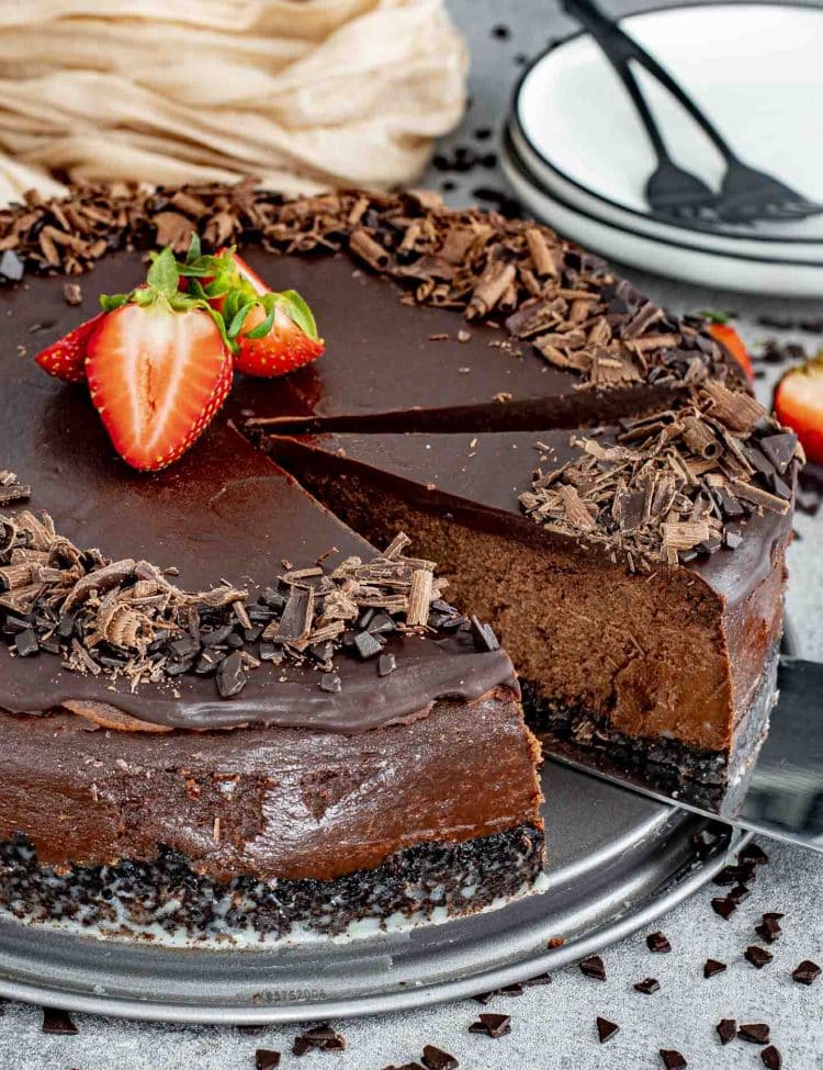A rich and creamy chocolate cheesecake with a glossy ganache topping, garnished with fresh strawberries and chocolate shavings. A slice is being lifted, revealing the velvety chocolate filling and a dark, crumbly chocolate cookie crust.