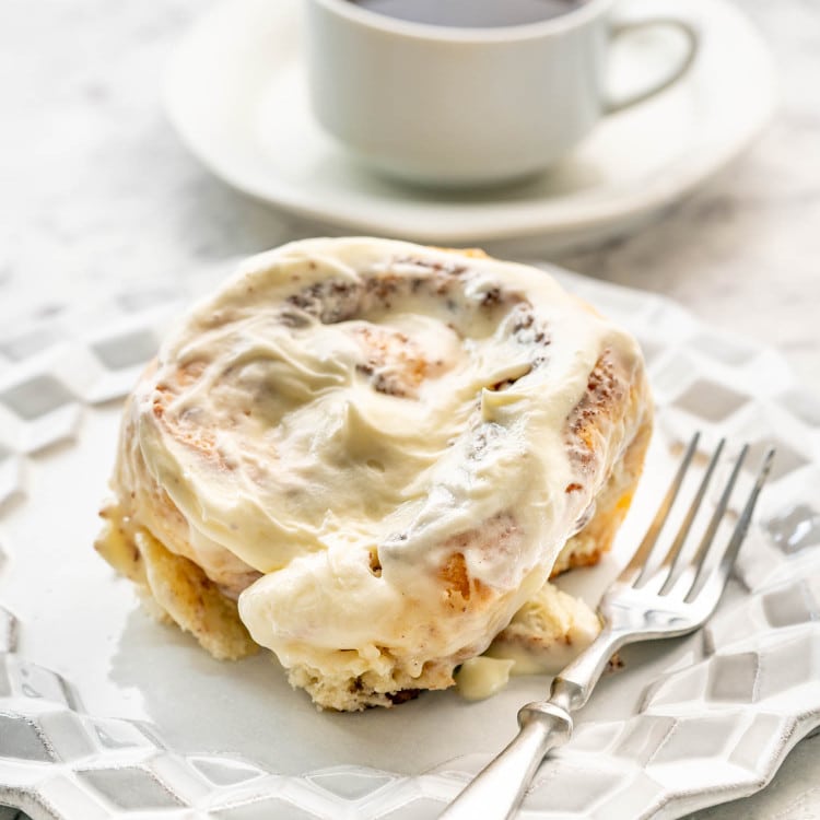 a cinnabon cinnamon roll on a white dessert plate with a cup of coffee in the background.