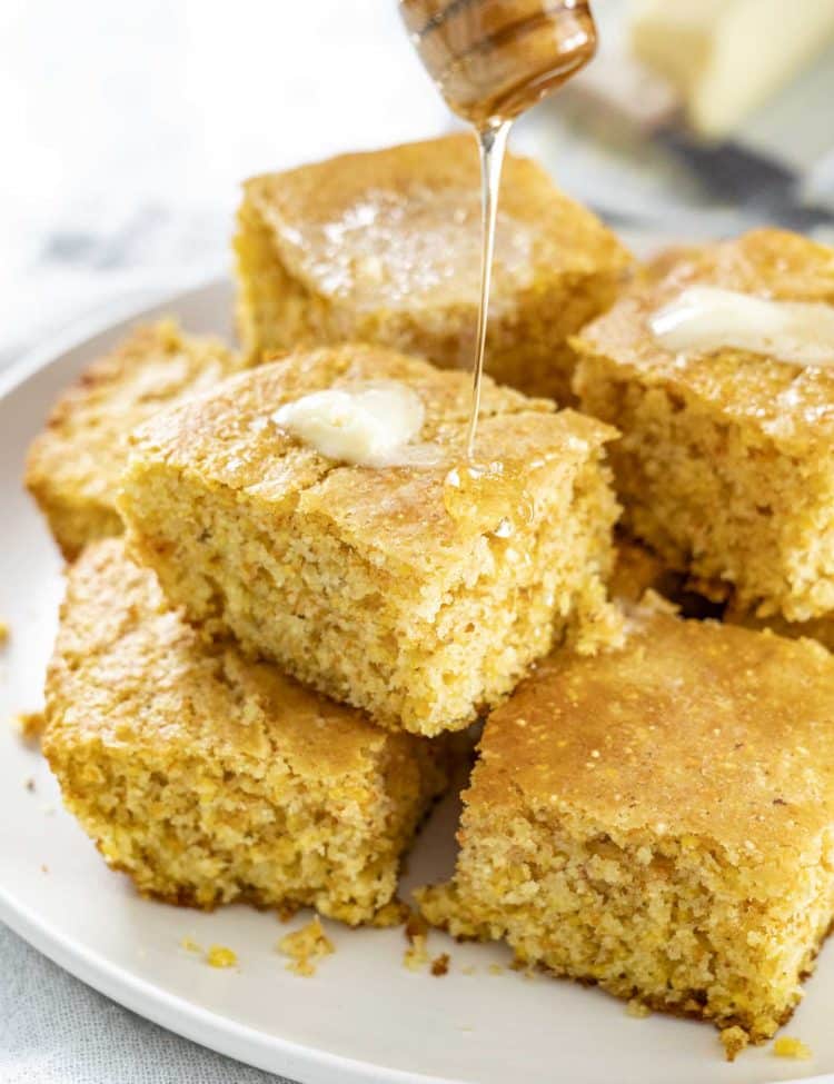 Golden squares of homemade cornbread topped with melting butter and drizzled with honey, served warm on a white plate.