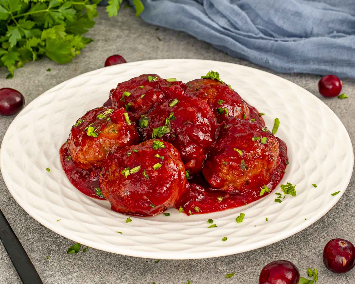 Plate of cranberry meatballs coated in vibrant cranberry sauce, surrounded by fresh cranberries and parsley leaves.