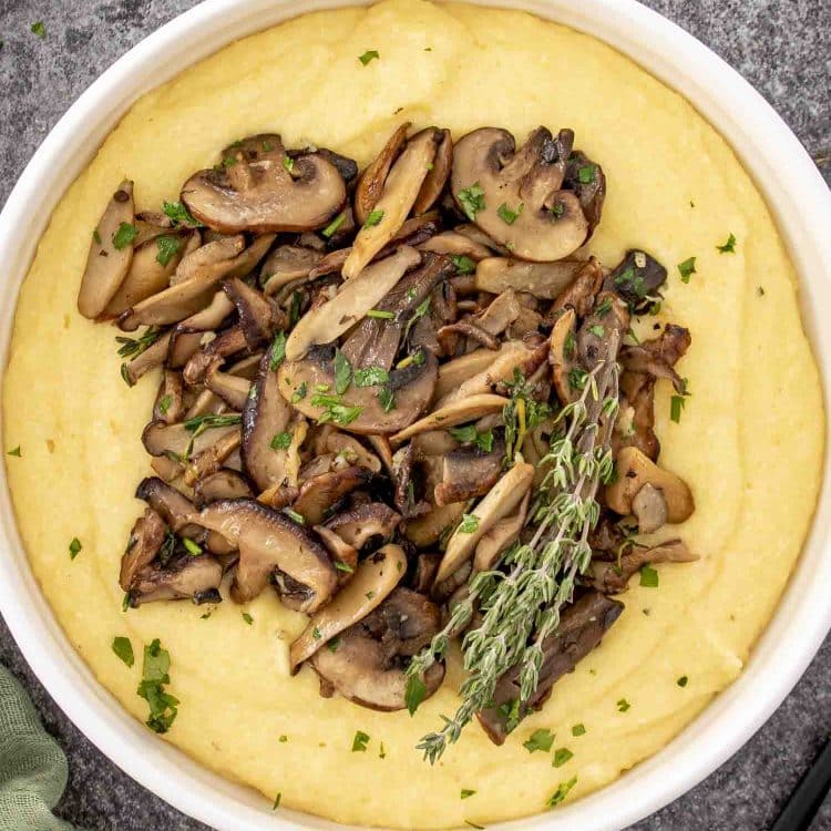 Overhead view of creamy Parmesan polenta with golden mushrooms and fresh thyme garnished with parsley in a round white bowl.