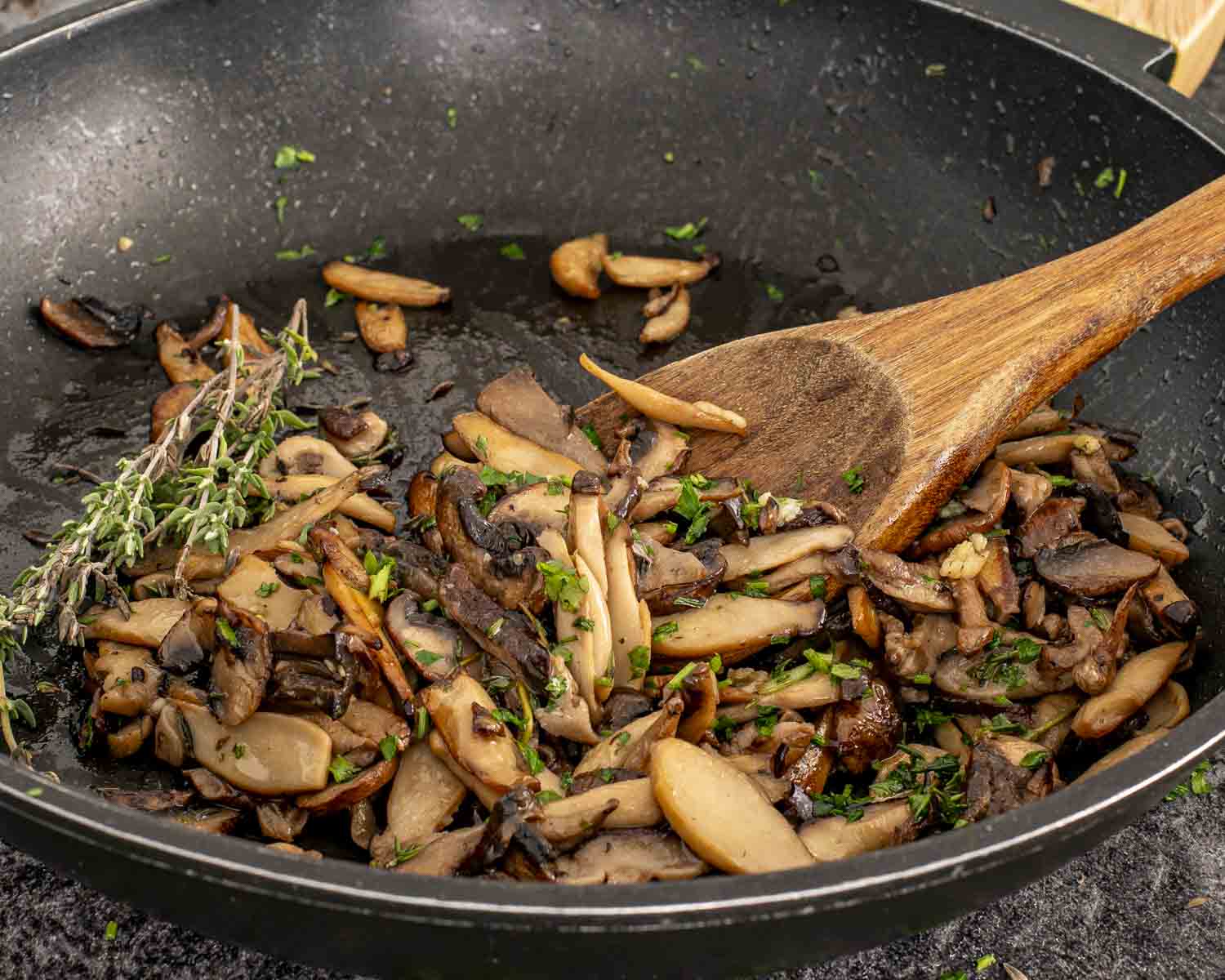 Mixed mushrooms sautéing in butter with garlic, parsley, and thyme inside a black skillet, stirred with a wooden spoon.