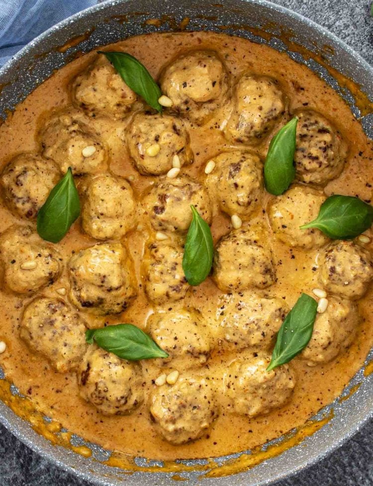 freshly made creamy sun dried tomato pesto meatballs in a skillet garnished with basil and pine nuts.