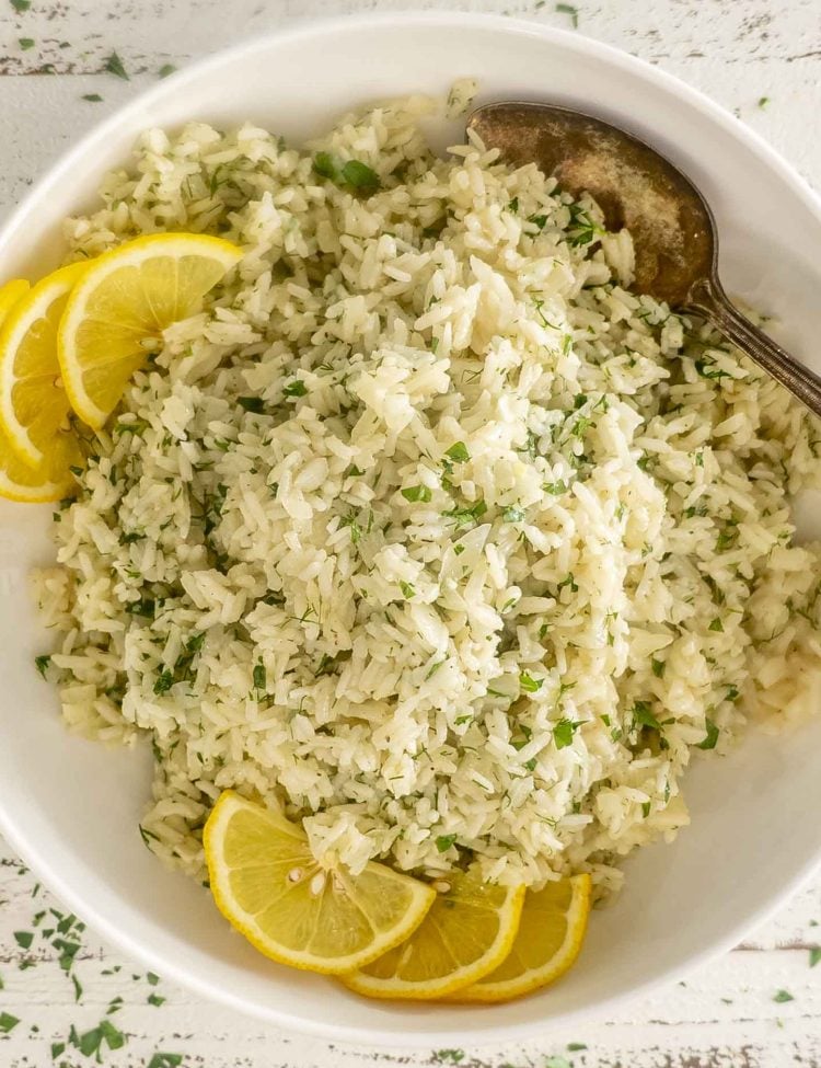 Overhead view of Greek rice in a white bowl with lemon wedges, herbs, and a vintage spoon.