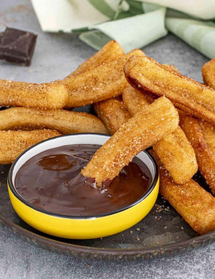 Cinnamon sugar churro dipped halfway into a bowl of rich melted chocolate, with more churros stacked beside it.