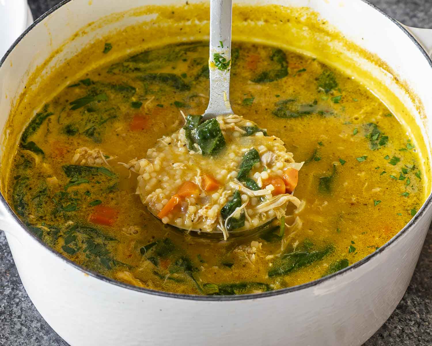 A ladle scooping Italian Penicillin Soup from a white pot showing ditalini pasta, shredded chicken, spinach, and carrots in broth.