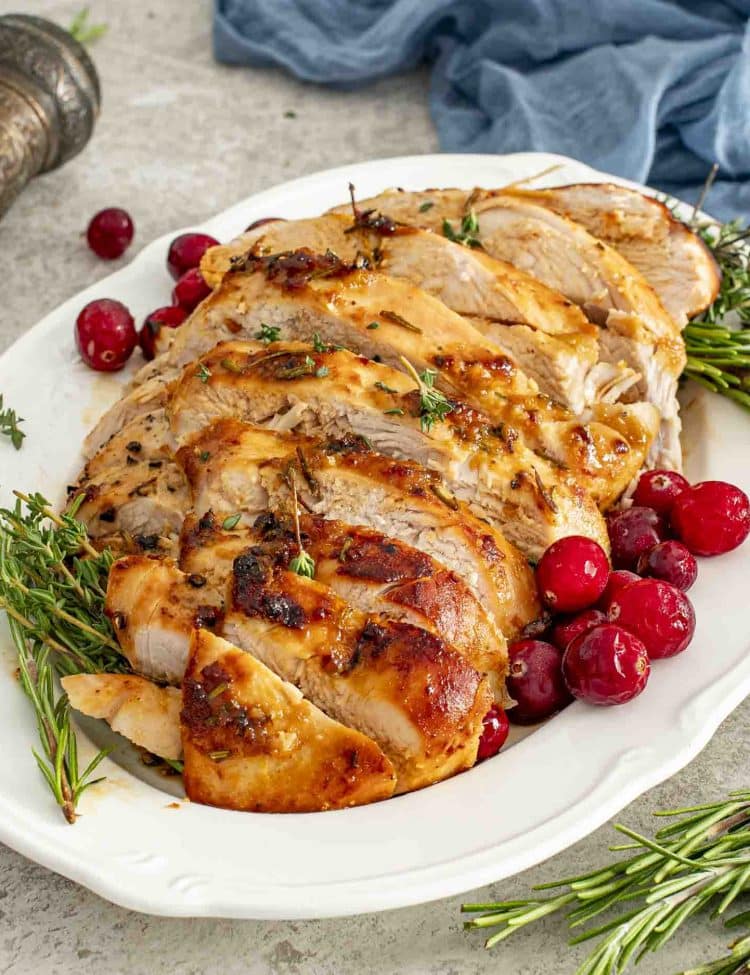 Juicy slices of Maple Dijon Glazed Turkey Breast arranged on a platter with rosemary, thyme, and bright red cranberries.
