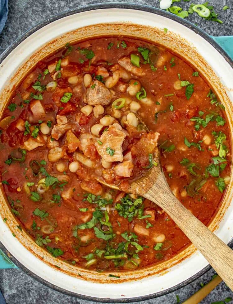 Bright red Mexican pork stew simmering in Dutch oven, filled with beans, tender pork, and chopped fresh herbs.