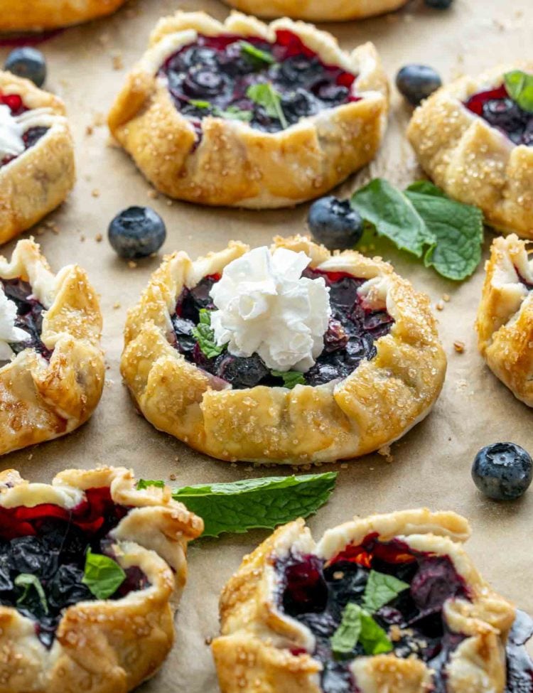 Freshly baked mini blueberry galette topped with whipped cream and mint leaves, surrounded by extra blueberries on parchment paper.