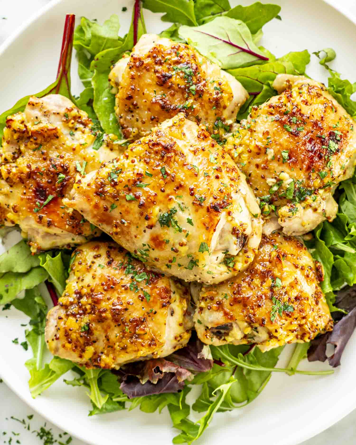 Close-up overhead of crispy chicken thighs on leafy greens, glazed and garnished with fresh chopped parsley.