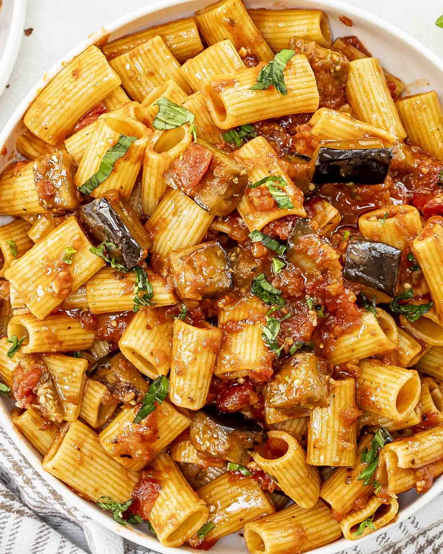 A white bowl of Pasta alla Norma featuring rigatoni in a rich tomato sauce with tender eggplant chunks, garnished with fresh basil.