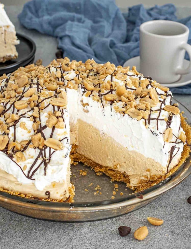 Whole peanut butter pie topped with whipped cream, chocolate drizzle, and peanuts with one slice removed for serving.