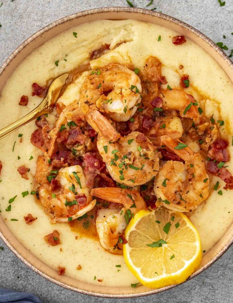 A bowl of creamy grits topped with juicy shrimp, crispy bacon, and fresh parsley, garnished with a lemon slice.