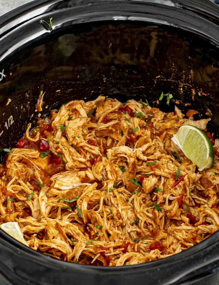 Shredded Slow Cooker Mexican Chicken in a black slow cooker, garnished with fresh cilantro and lime wedges.
