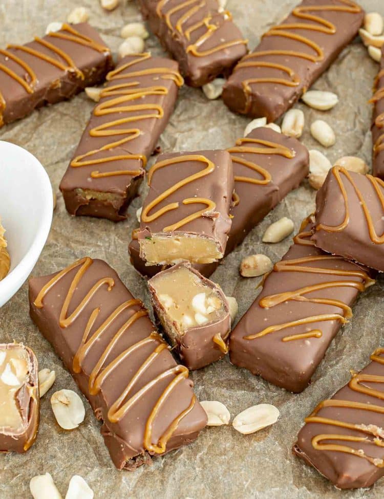 Rows of homemade Snickers Bites on parchment, coated in chocolate and caramel drizzle, with peanuts scattered around for texture.