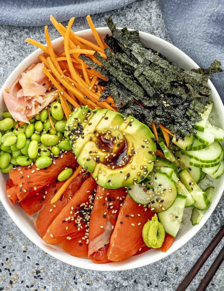 A sushi bowl filled with sushi rice topped with sliced salmon, avocado drizzled with soy sauce, cucumber, shredded carrots, edamame, and thin strips of nori.