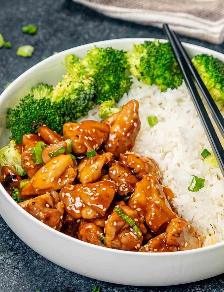 teriyaki chicken in a white bowl with some cooked rice and steamed broccoli.