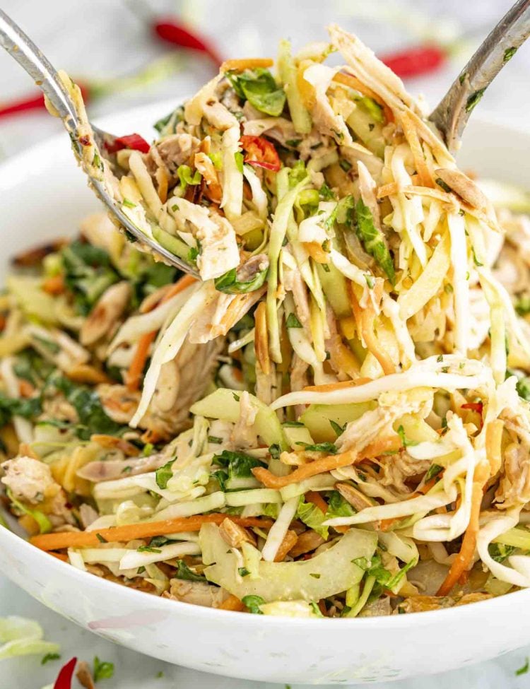 Close-up of Thai Chicken Salad with shredded cabbage, papaya, carrots, and herbs being tossed with forks.