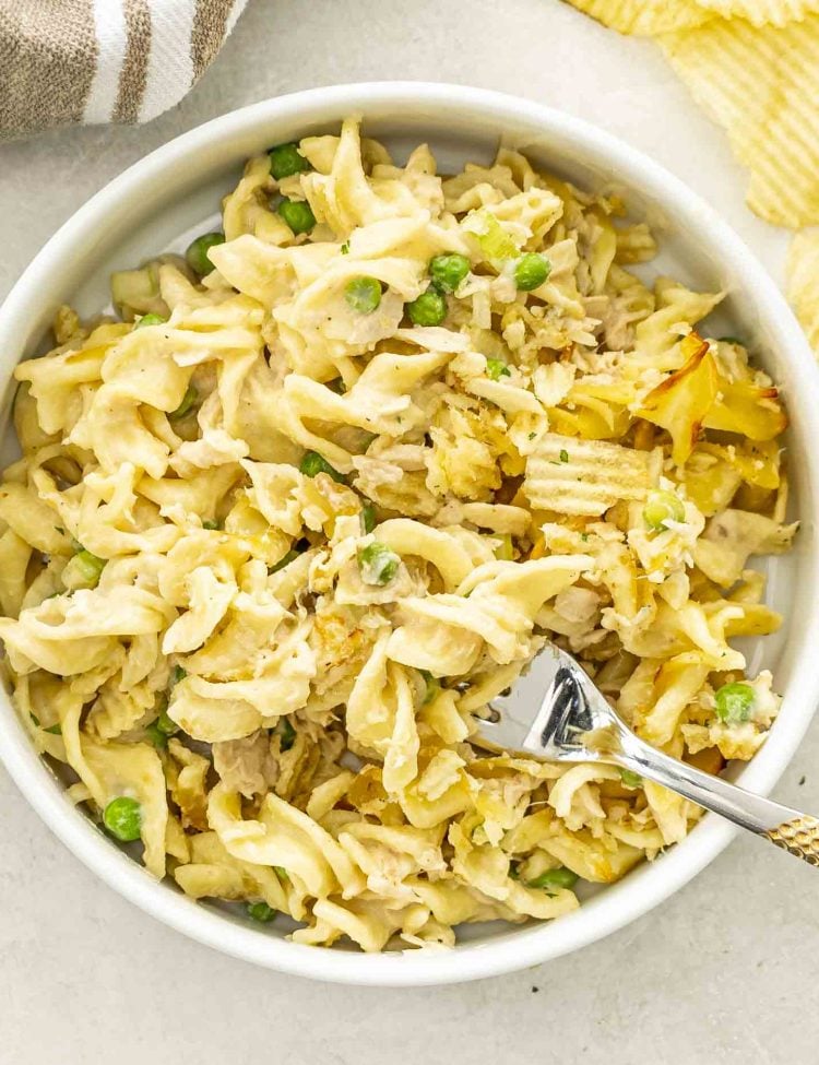 Overhead view of tuna noodle casserole in a white bowl, showcasing creamy noodles, tuna, peas, and golden topping.