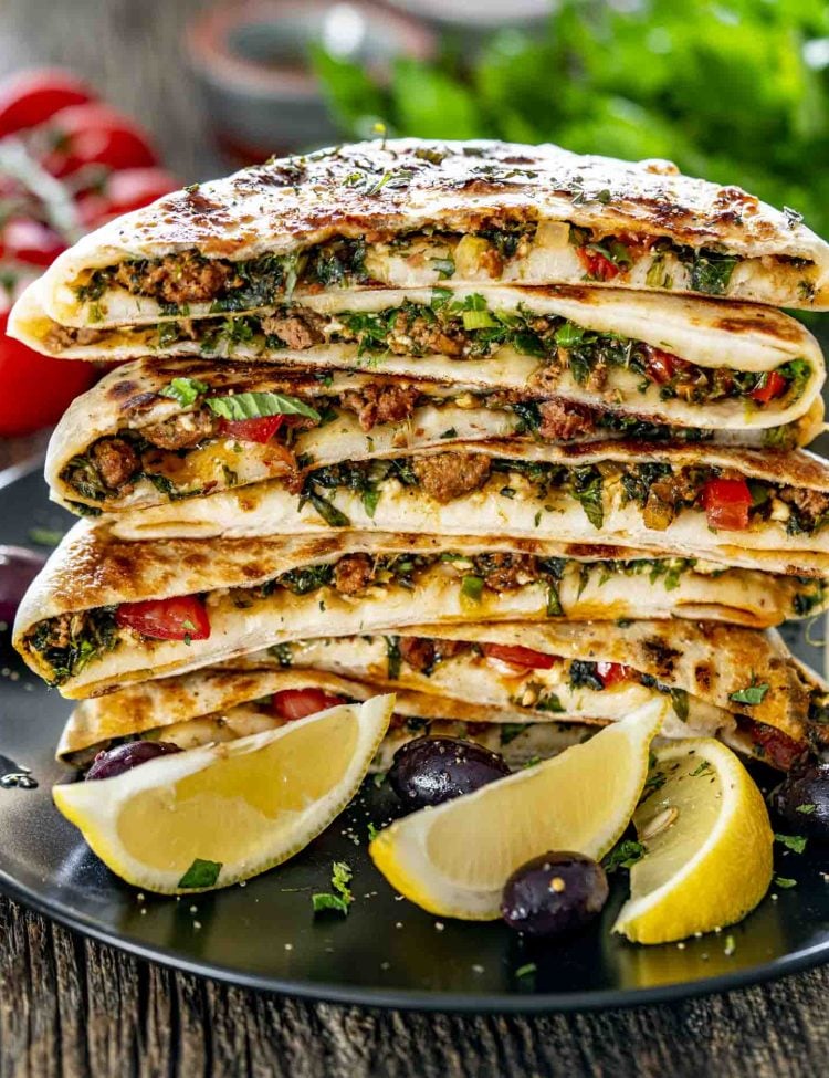 Stacked slices of Turkish Gozleme with lamb, herbs, and feta on a black plate with lemon wedges.